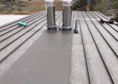 Two metal flue pipes with protective covers on a corrugated metal roof with installation tools nearby.