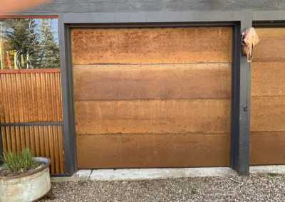 Rusty metal garage door beside a wooden fence and a potted plant.