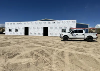 A white pickup truck parked in front of a building under construction wrapped in tyvek house wrap, with a clear sky and dirt ground.