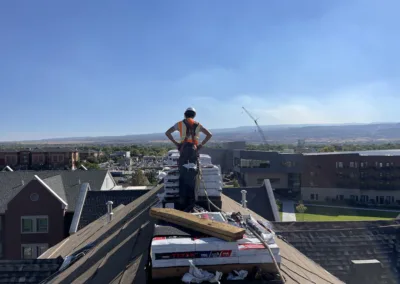 Construction worker standing on a sloped rooftop with building materials, overlooking a residential area.