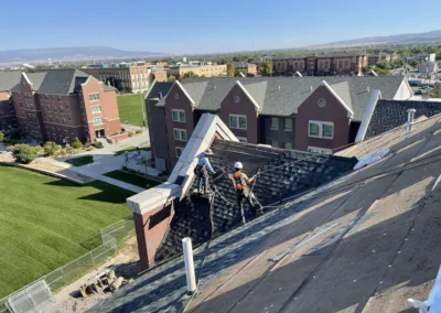 Workers installing roofing materials on a steep slope with buildings and a landscape in the background.