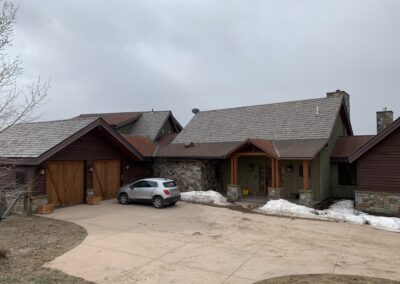 A large house with a brown shingle roof, stone and wood siding, a garage, and a white suv parked in the driveway under an overcast sky. snow is visible on the ground.