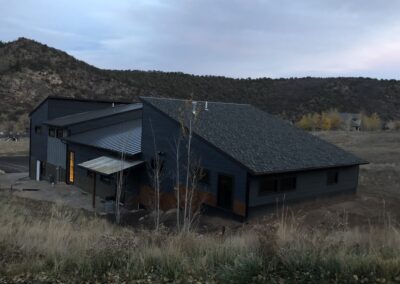 A modern house with a dark sloped roof and gray siding located in a rural area with hills in the background during dusk.