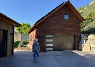 A person in a hat stands on a paved driveway facing a brown, modern garage with a sloped roof and solar panels, surrounded by greenery and rocks.